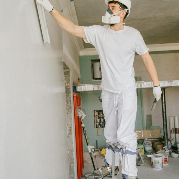 Construction worker on stilts painting a wall indoors, wearing protective gear and a hard hat.